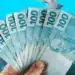 Mão segurando várias notas de 100 reais abertas em leque sobre fundo azul.