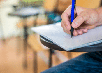 Estudante escrevendo prova em carteira escolar durante exame presencial.