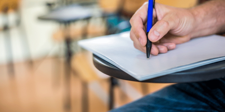 Estudante escrevendo prova em carteira escolar durante exame presencial.