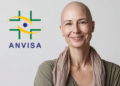 Mulher adulta careca sorri ao lado do logo da ANVISA em fundo claro.