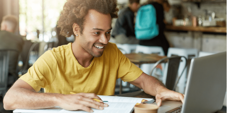 Jovem sorridente estuda em cafeteria com notebook e livros.