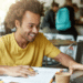 Jovem sorridente estuda em cafeteria com notebook e livros.