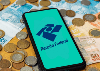 Logo da Receita Federal em destaque no celular, com notas e moedas de real ao redor.