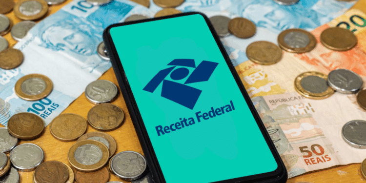 Logo da Receita Federal em destaque no celular, com notas e moedas de real ao redor.