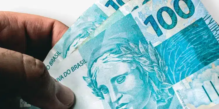Mão segurando duas notas de 100 reais com arte da República em destaque.