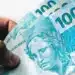 Mão segurando duas notas de 100 reais com arte da República em destaque.