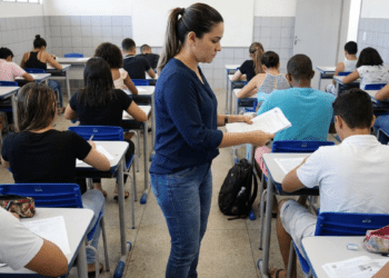 Fiscal de concursos entrega provas em sala com estudantes em exame.