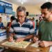 Dois homens preenchem bilhetes de loteria em uma casa lotérica com placas da Caixa e do concurso 6082 ao fundo