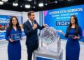 Um apresentador e duas assistentes de palco conduzem sorteio da Loteria dos Sonhos com globos e painel digital em estúdio iluminado