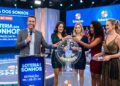 Apresentador sorridente com quatro mulheres sorteando bolinhas no globo da Loteria dos Sonhos, palco azul iluminado