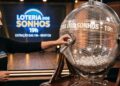 Mulher sorteando bola número 42 na Loteria dos Sonhos das 19h com globo transparente e fundo do palco