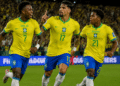 Três jogadores da Seleção Brasileira comemoram gol vestindo uniformes amarelos com nomes Viní Jr., Paquetá e Endrick.