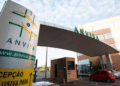 Entrada da sede da Anvisa com placa, estrutura moderna, carros e céu azul ao fundo.