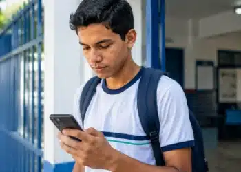 Estudante com mochila consultando o celular na entrada da escola, representando o acesso à conta do estudante pelo aplicativo Caixa Tem.