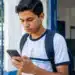 Estudante com mochila consultando o celular na entrada da escola, representando o acesso à conta do estudante pelo aplicativo Caixa Tem.