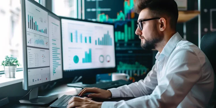 Analista de sistemas trabalhando em computador com múltiplos monitores exibindo gráficos e dados tecnológicos.