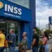 Fila de segurados em frente a uma agência do INSS, com fachada azul em destaque, relacionada a projeto sobre aposentadoria maior.