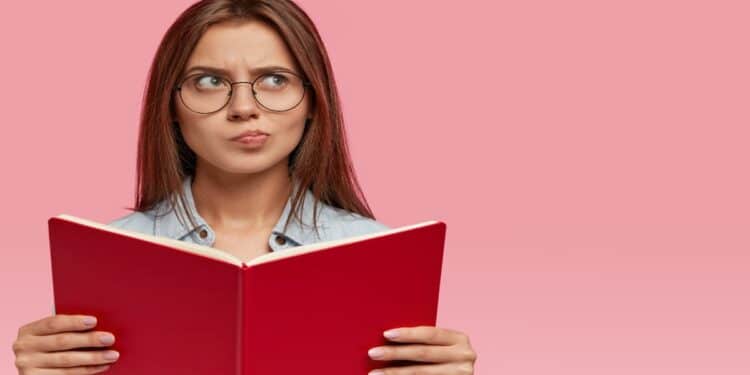Jovem de óculos segura livro aberto com expressão de dúvida, referência ao erro comum na grafia da palavra através.