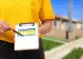 Imagem ilustrativa de atendente de colete amarelo segurando prancheta com a marca do Cadastro Único diante de casas de conjunto habitacional.