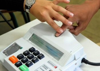 Imagem ilustrativa de eleitor pressionando o dedo no leitor biométrico acoplado ao terminal usado pelo mesário, equipamento com teclado numérico e botões de correção e confirmação