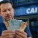 Trabalhador segura notas de real em frente à Caixa, representando o cálculo do valor do lucro do FGTS.