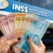 Idosa segurando cédulas de real em frente a uma agência do INSS, sobre o 13º salário do INSS em novembro