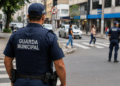 Guarda municipal fardado acompanha o movimento de veículos e pedestres em uma rua da cidade.