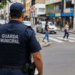 Guarda municipal fardado acompanha o movimento de veículos e pedestres em uma rua da cidade.