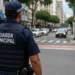 Guarda municipal uniformizado observa o trânsito e a movimentação de pedestres em uma rua urbana.