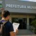 Pessoa segurando documento de concurso público em frente à Prefeitura Municipal de Bálsamo-SP.