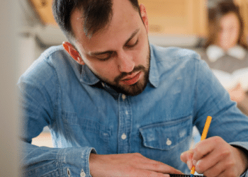 Homem jovem de barba, camisa jeans azul, escrevendo com lápis enquanto mulher lê ao fundo.