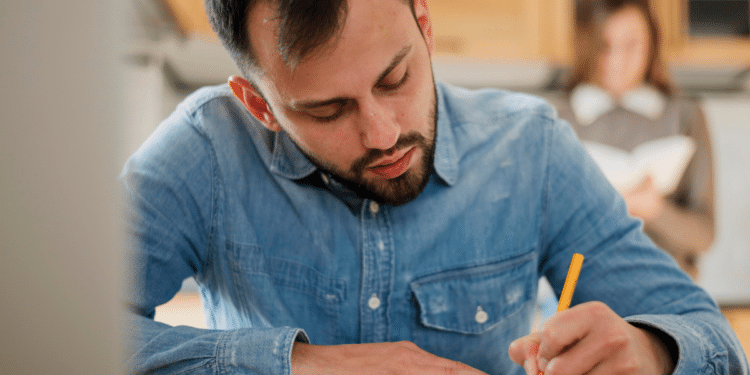 Homem jovem de barba, camisa jeans azul, escrevendo com lápis enquanto mulher lê ao fundo.