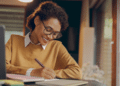 Mulher jovem sorrindo, escrevendo em caderno, usando suéter mostarda, óculos e sentada em mesa com notebook.