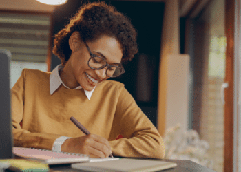 Mulher jovem sorrindo, escrevendo em caderno, usando suéter mostarda, óculos e sentada em mesa com notebook.