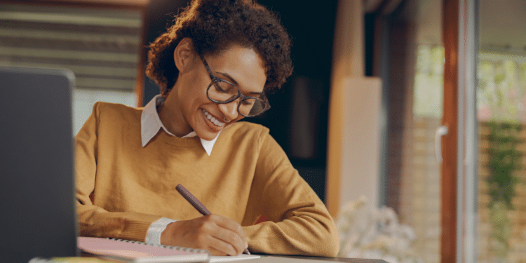 Mulher jovem sorrindo, escrevendo em caderno, usando suéter mostarda, óculos e sentada em mesa com notebook.