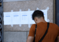 Pessoa consultando lista de aprovados em concurso do Ministério Público fixada na parede.