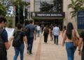 Candidatos caminham em direção à prefeitura com mochilas e documentos de concurso público.