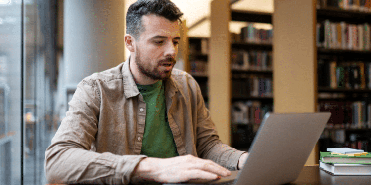 Homem estudando para concurso público utilizando notebook em uma biblioteca.