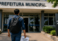 Pessoa caminhando em direção à Prefeitura Municipal segurando documento de concurso público em área externa.