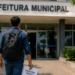 Pessoa caminhando em direção à Prefeitura Municipal segurando documento de concurso público em área externa.
