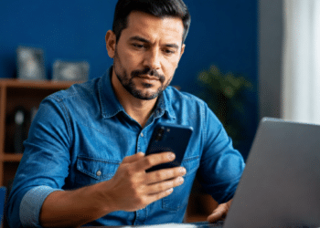 Homem consulta pelo celular o terceiro lote da restituição do Imposto de Renda enquanto utiliza um notebook.