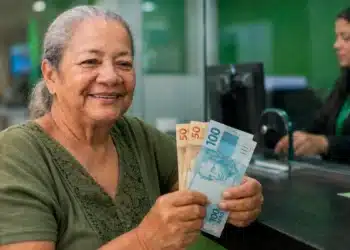 Aposentada segura notas de real em uma agência bancária, representando a devolução de R$ 547 milhões pelo INSS às vítimas de descontos indevidos.