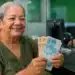 Aposentada segura notas de real em uma agência bancária, representando a devolução de R$ 547 milhões pelo INSS às vítimas de descontos indevidos.