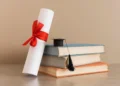 Diploma enrolado com laço vermelho ao lado de livros e capelo de formatura.