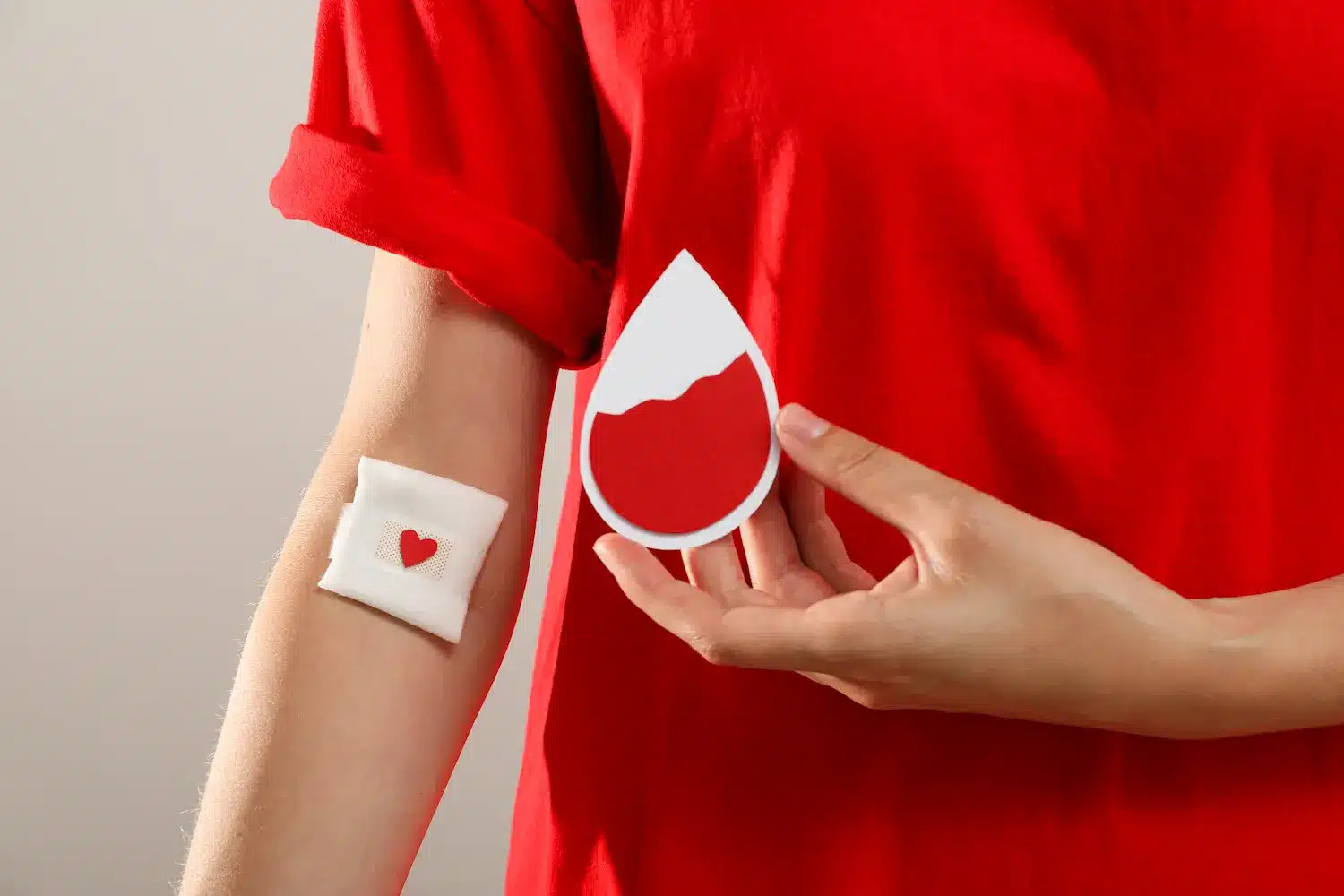 Doação de sangue ajuda a manter estoques dos hemocentros Pessoa com curativo no braço após doação de sangue segurando símbolo de gota vermelha.