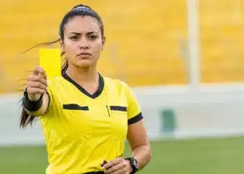 Mulher em campo segurando cartão amarelo durante partida esportiva, representando o feminino correto de árbitro.