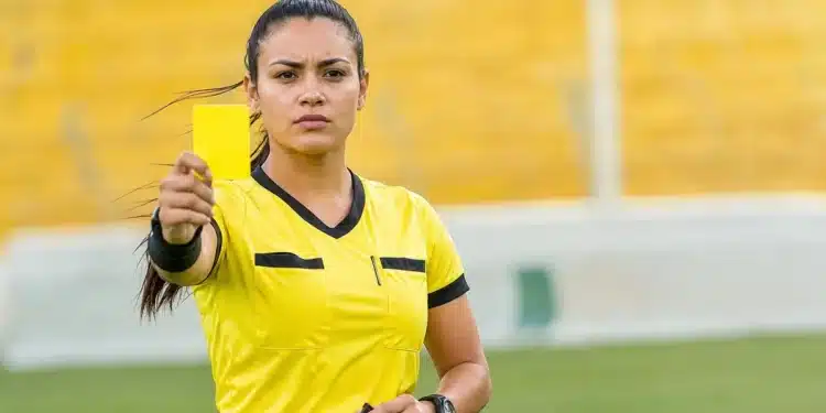 Mulher em campo segurando cartão amarelo durante partida esportiva, representando o feminino correto de árbitro.