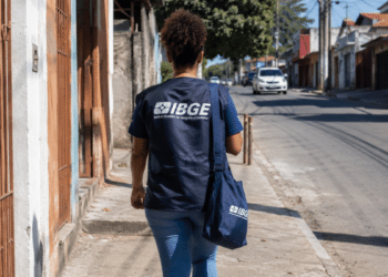 Agente do IBGE uniformizada caminha por rua residencial durante trabalho de campo.