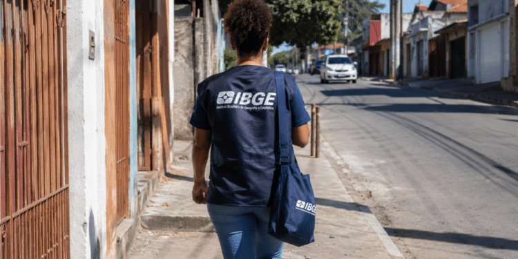 Agente do IBGE uniformizada caminha por rua residencial durante trabalho de campo.