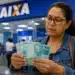 Mulher conferindo cédulas de R$ 100 em uma agência da Caixa, representando a liberação de valores esquecidos do PIS/Pasep.